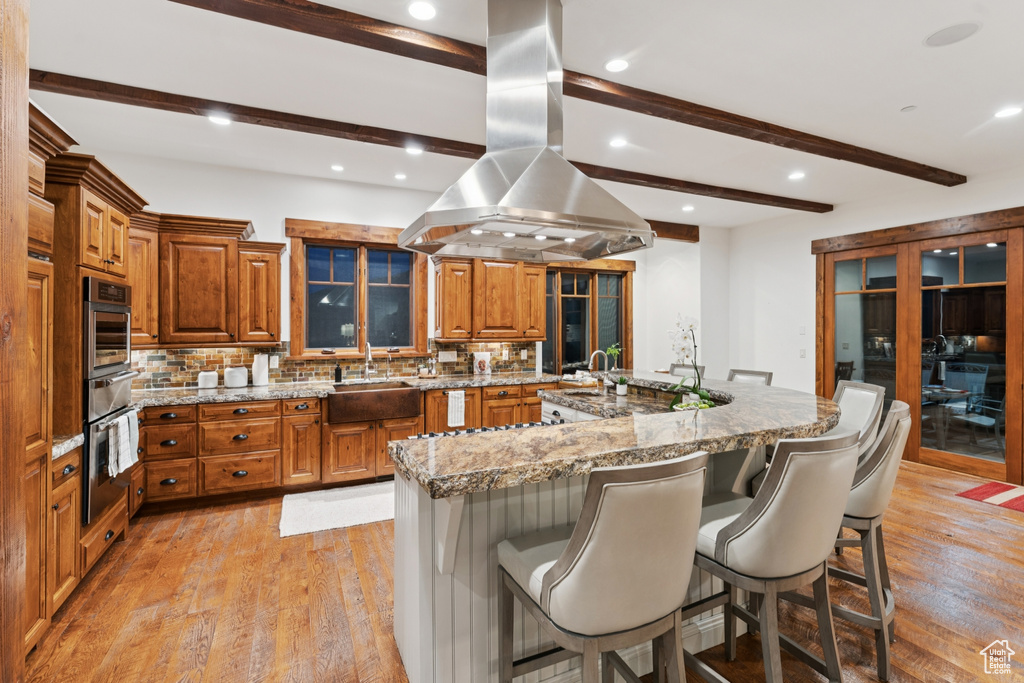 Kitchen with island range hood, oven, beamed ceiling, brown cabinets, and backsplash