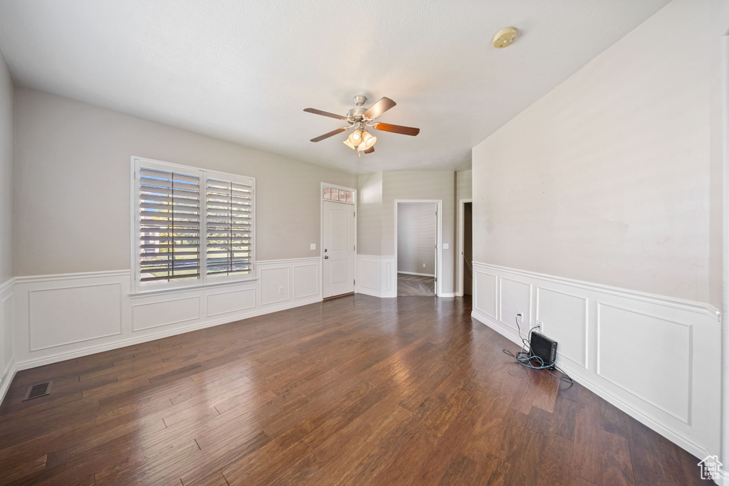 Empty room with a ceiling fan, a smoke detector, a wainscoted wall, dark wood finished floors, and a decorative wall