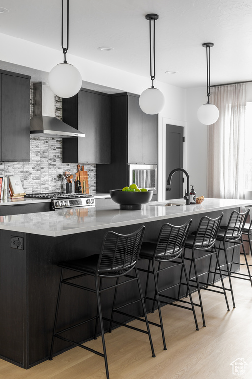 Kitchen featuring wall chimney exhaust hood, dark cabinets, light wood-style flooring, and a kitchen bar