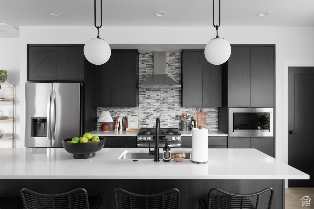 Kitchen featuring appliances with stainless steel finishes, wall chimney exhaust hood, a kitchen bar, light countertops, and pendant lighting