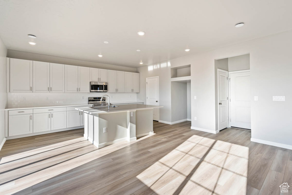 Kitchen featuring stainless steel appliances, an island with sink, light countertops, white cabinets, and light wood-style floors