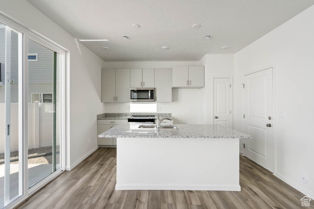 Kitchen featuring stainless steel microwave, light wood-style floors, light stone countertops, an island with sink, and a textured ceiling