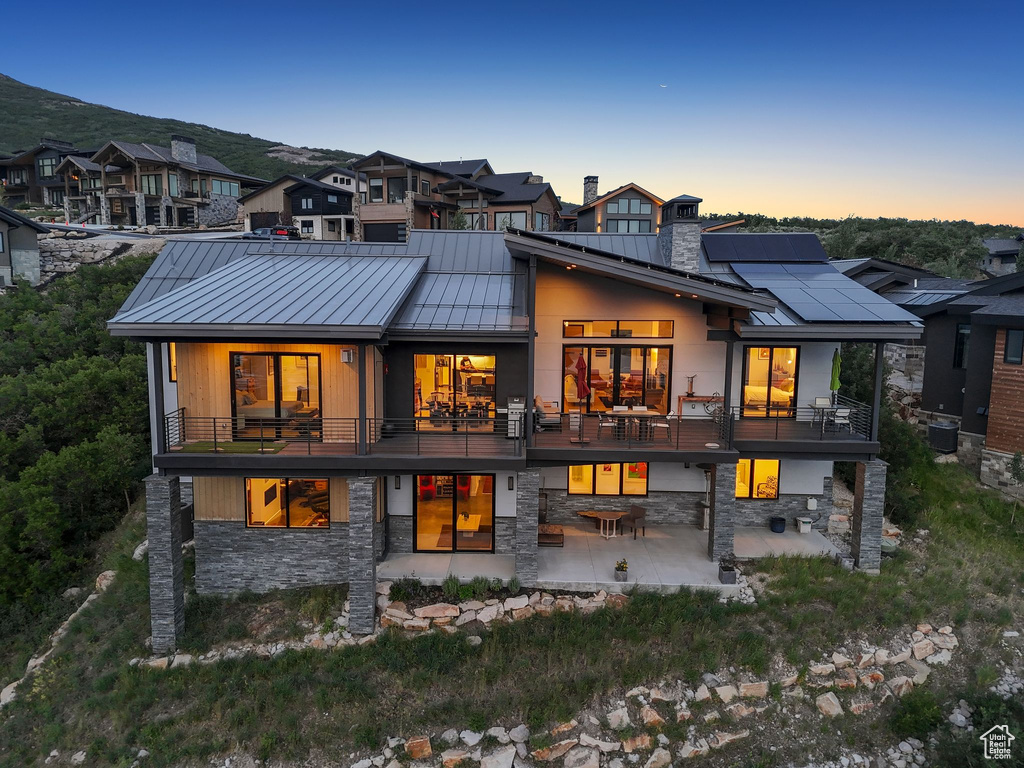 Back of property at dusk featuring a standing seam roof, a patio, stone siding, and a balcony
