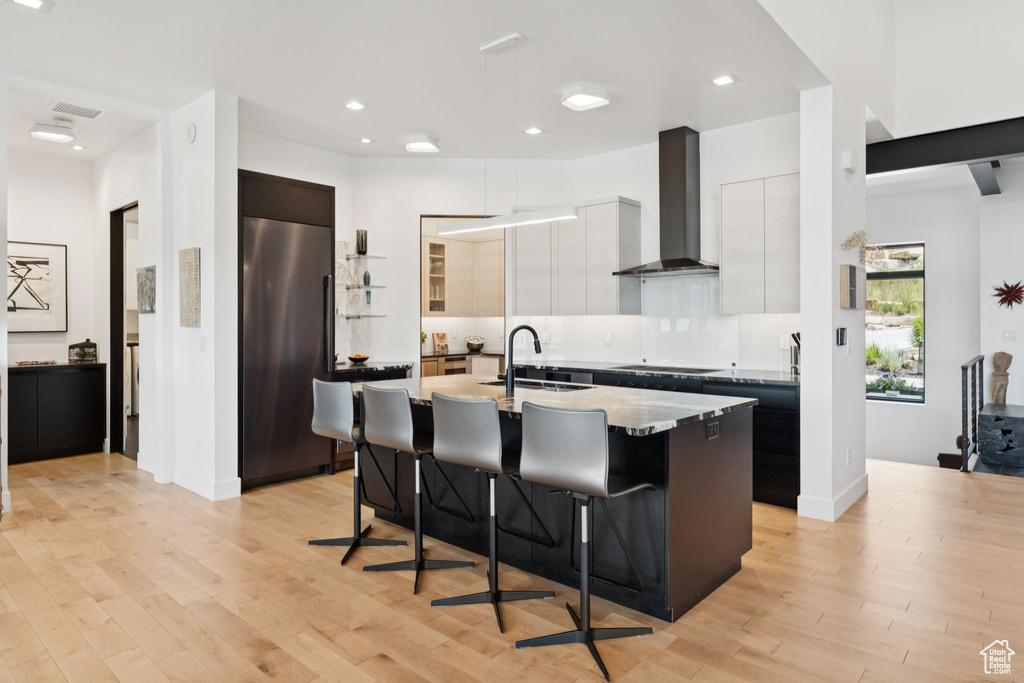 Kitchen with wall chimney range hood, high end refrigerator, modern cabinets, a breakfast bar area, and a kitchen island with sink