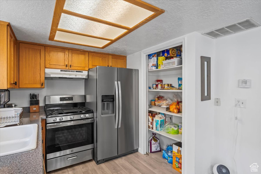 Kitchen with appliances with stainless steel finishes, under cabinet range hood, a textured ceiling, light wood-style floors, and brown cabinets