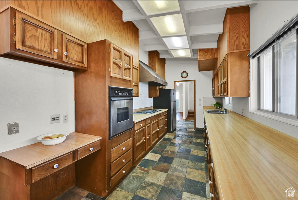 Kitchen featuring stainless steel appliances, dark stone finish floors, brown cabinets, exhaust hood, and coffered ceiling