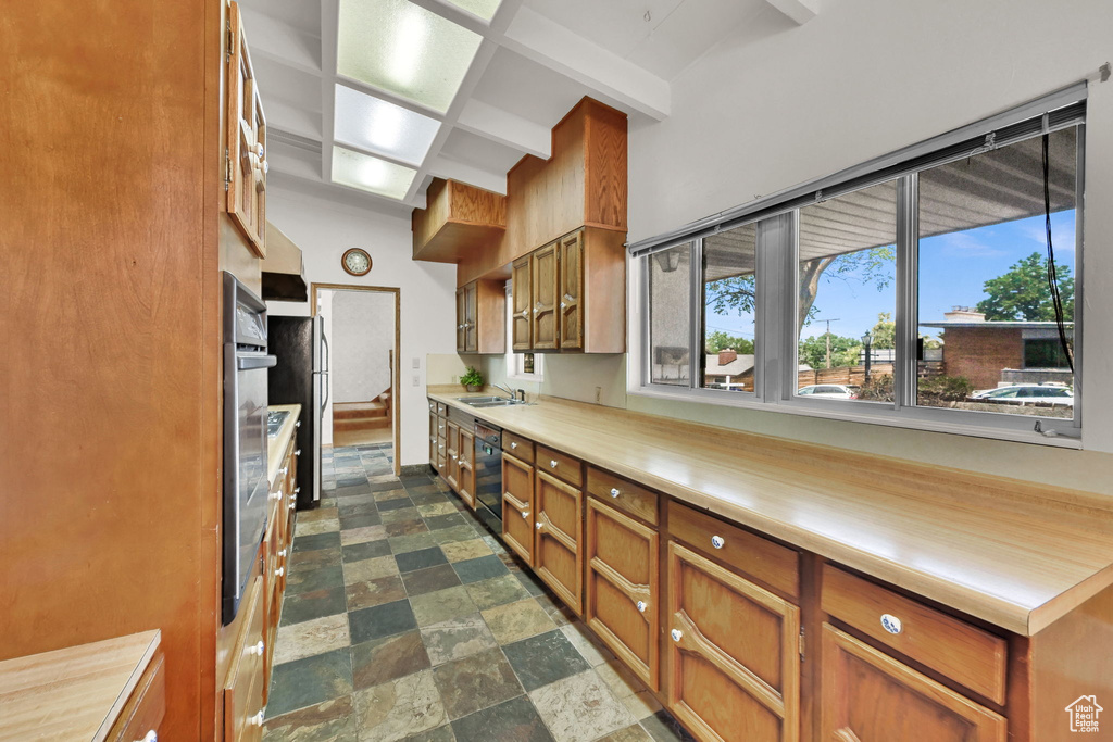 Kitchen with brown cabinetry, light countertops, stone finish floors, beam ceiling, and black dishwasher
