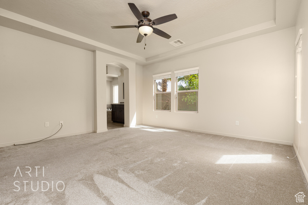 Carpeted empty room featuring a raised ceiling, arched walkways, and ceiling fan