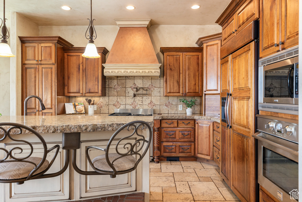 Kitchen featuring custom range hood, built in appliances, brown cabinetry, recessed lighting, and stone tile flooring