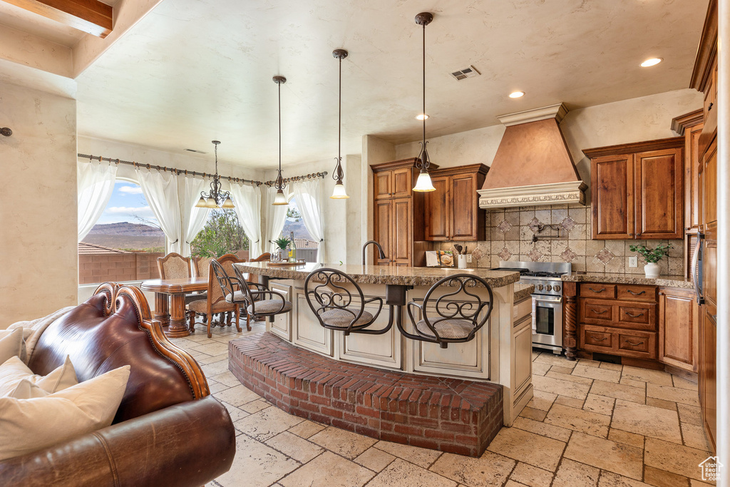 Kitchen featuring custom exhaust hood, stainless steel range, stone tile floors, brown cabinetry, and a kitchen breakfast bar