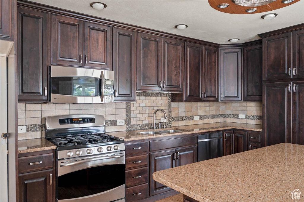 Kitchen featuring stainless steel appliances, dark brown cabinets, backsplash, and light stone countertops