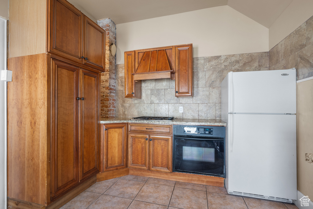 Kitchen with freestanding refrigerator, black oven, custom exhaust hood, brown cabinetry, and vaulted ceiling