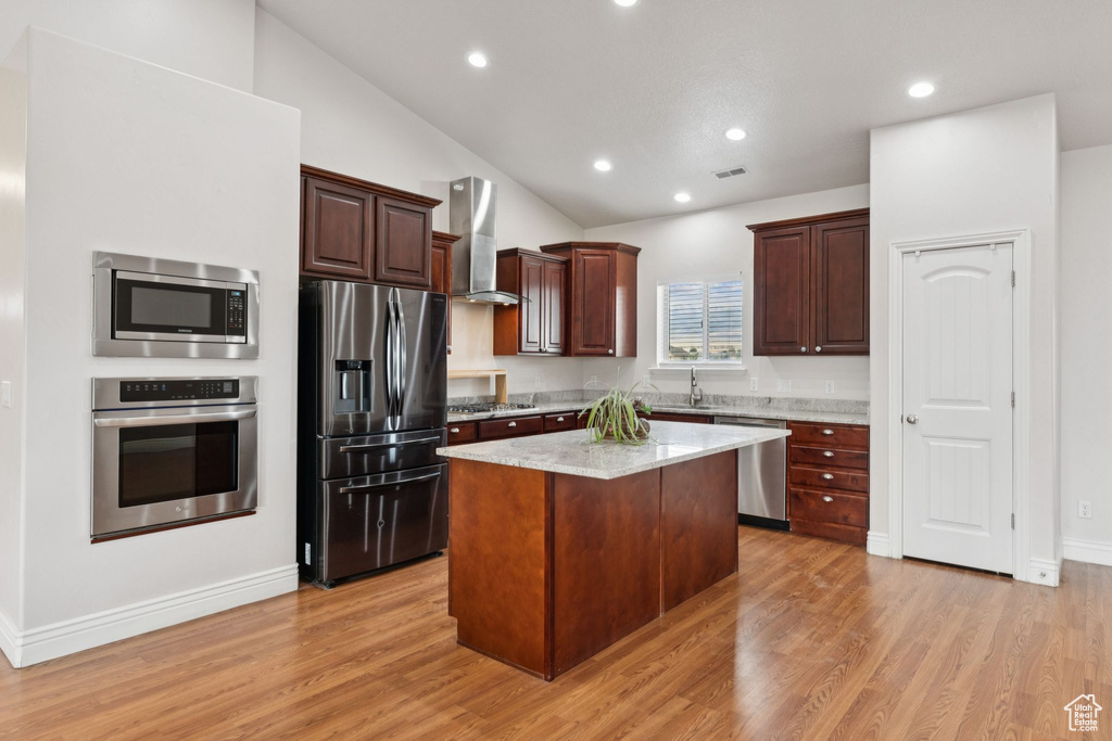 Kitchen featuring appliances with stainless steel finishes, wall chimney exhaust hood, vaulted ceiling, a center island, and light wood-style flooring