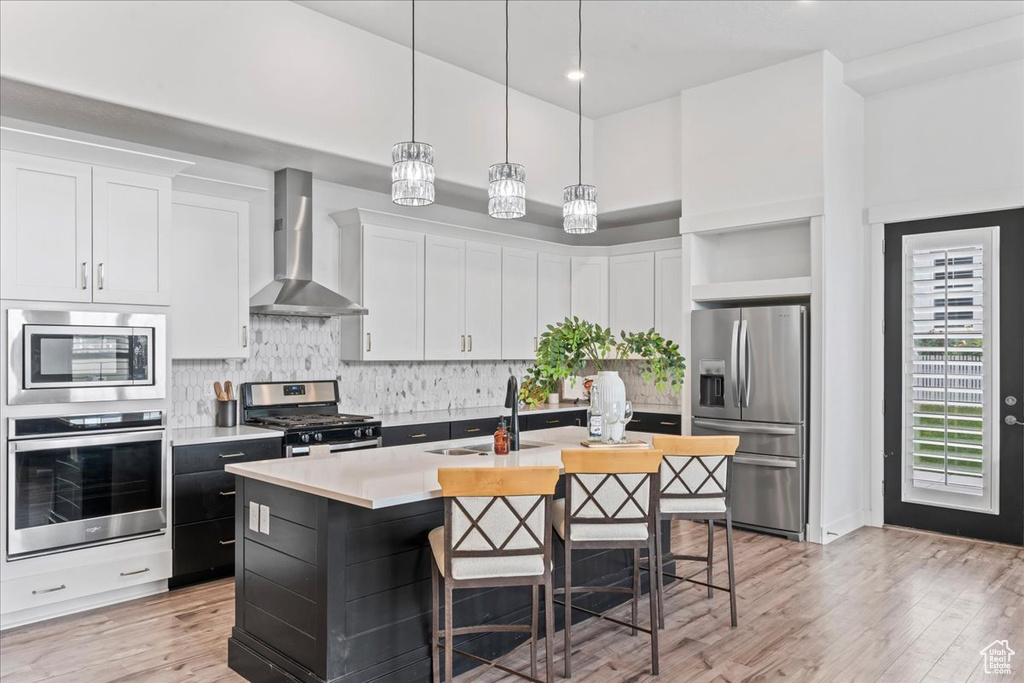 Kitchen featuring stainless steel appliances, wall chimney range hood, light countertops, a center island with sink, and light wood-type flooring