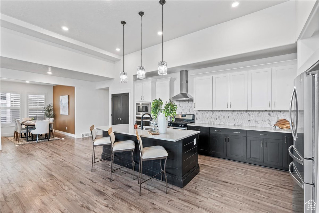 Kitchen with wall chimney exhaust hood, appliances with stainless steel finishes, a breakfast bar, light countertops, and light wood-style flooring