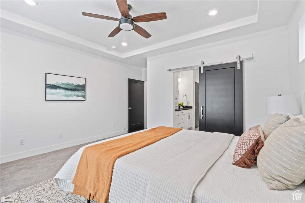 Carpeted bedroom with a barn door, a tray ceiling, recessed lighting, ensuite bath, and a ceiling fan