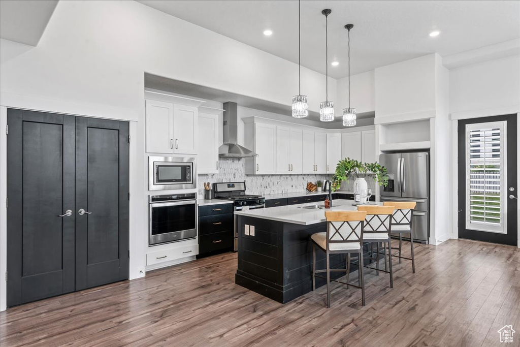 Kitchen with stainless steel appliances, wall chimney exhaust hood, white cabinets, a center island with sink, and tasteful backsplash