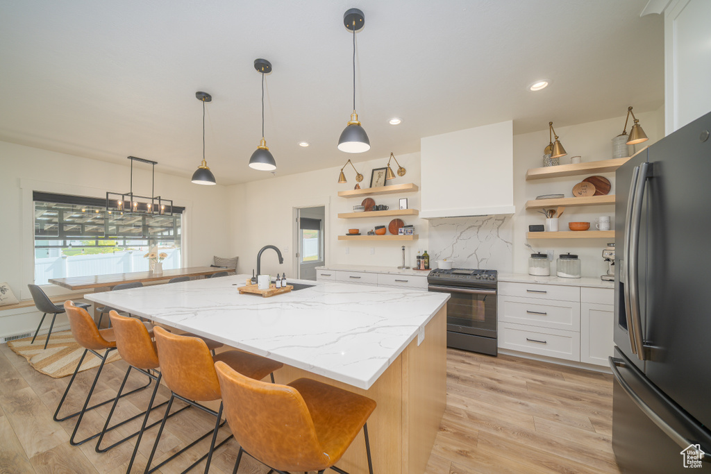 Kitchen with open shelves, appliances with stainless steel finishes, light wood finished floors, a center island with sink, and recessed lighting