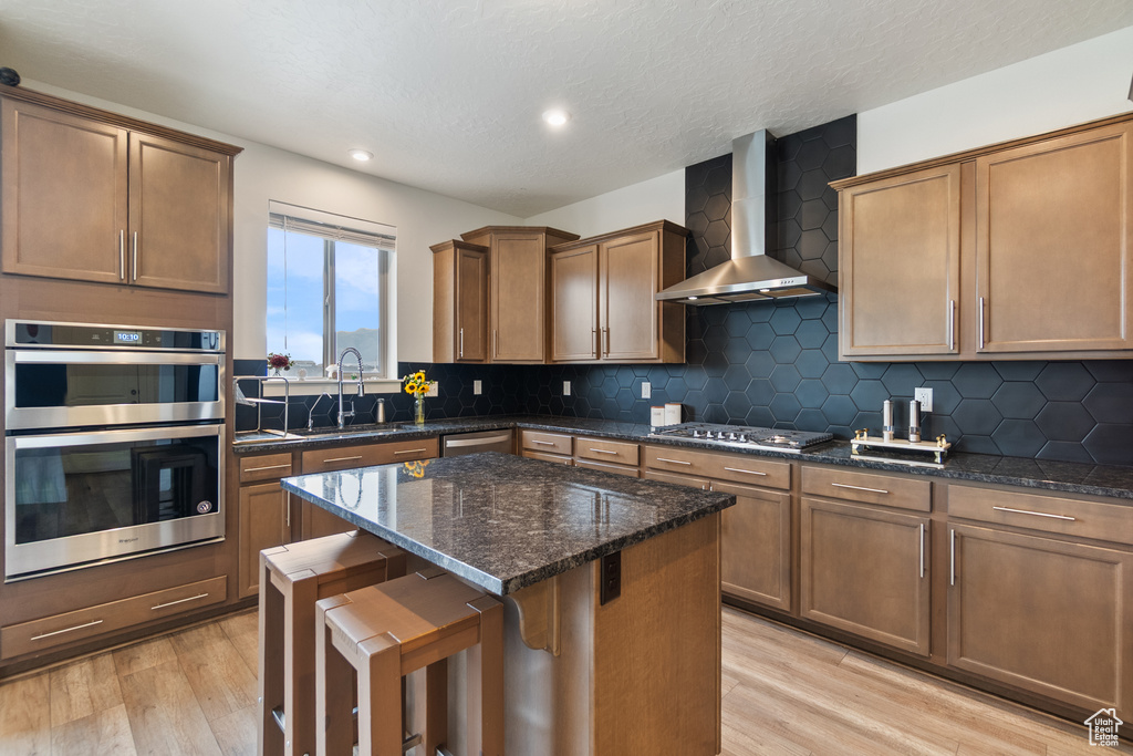 Kitchen featuring wall chimney exhaust hood, stainless steel appliances, light wood-type flooring, a kitchen island, and dark stone countertops