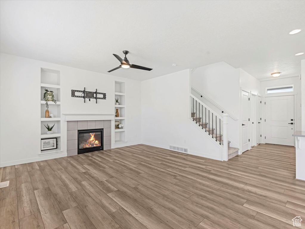 Unfurnished living room featuring stairway, light wood-type flooring, built in shelves, a ceiling fan, and a tile fireplace