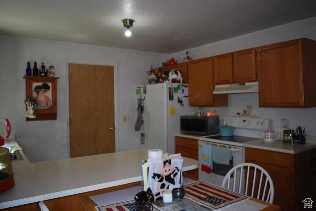 Kitchen featuring white appliances, under cabinet range hood, brown cabinetry, light countertops, and a peninsula
