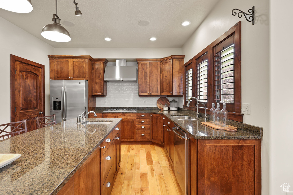 Kitchen with appliances with stainless steel finishes, wall chimney range hood, light wood-type flooring, brown cabinetry, and recessed lighting
