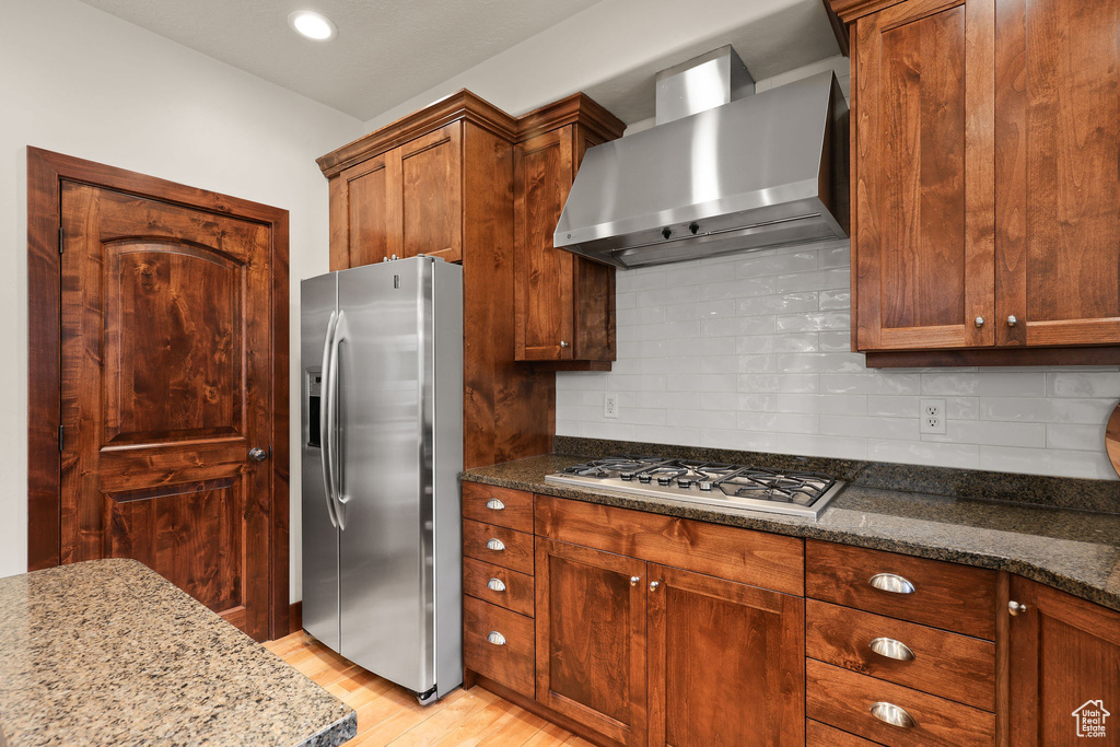 Kitchen with appliances with stainless steel finishes, wall chimney range hood, light wood finished floors, dark stone countertops, and decorative backsplash