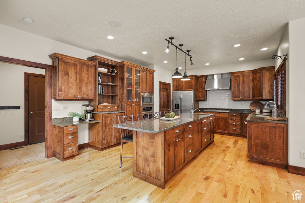 Kitchen featuring wall chimney exhaust hood, stainless steel appliances, an island with sink, recessed lighting, and light wood finished floors