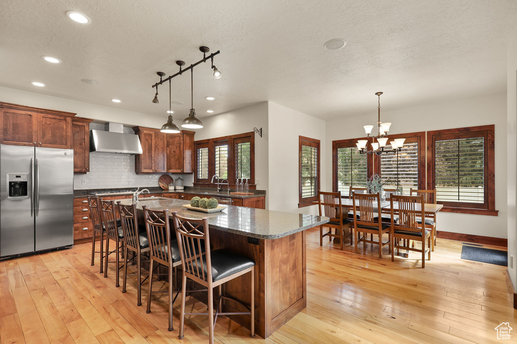 Kitchen with stainless steel refrigerator with ice dispenser, wall chimney exhaust hood, a textured ceiling, recessed lighting, and an island with sink