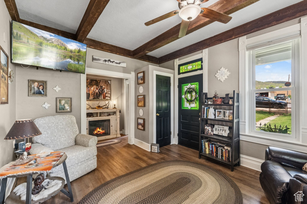 Living room featuring beamed ceiling, a glass covered fireplace, wood finished floors, and a ceiling fan