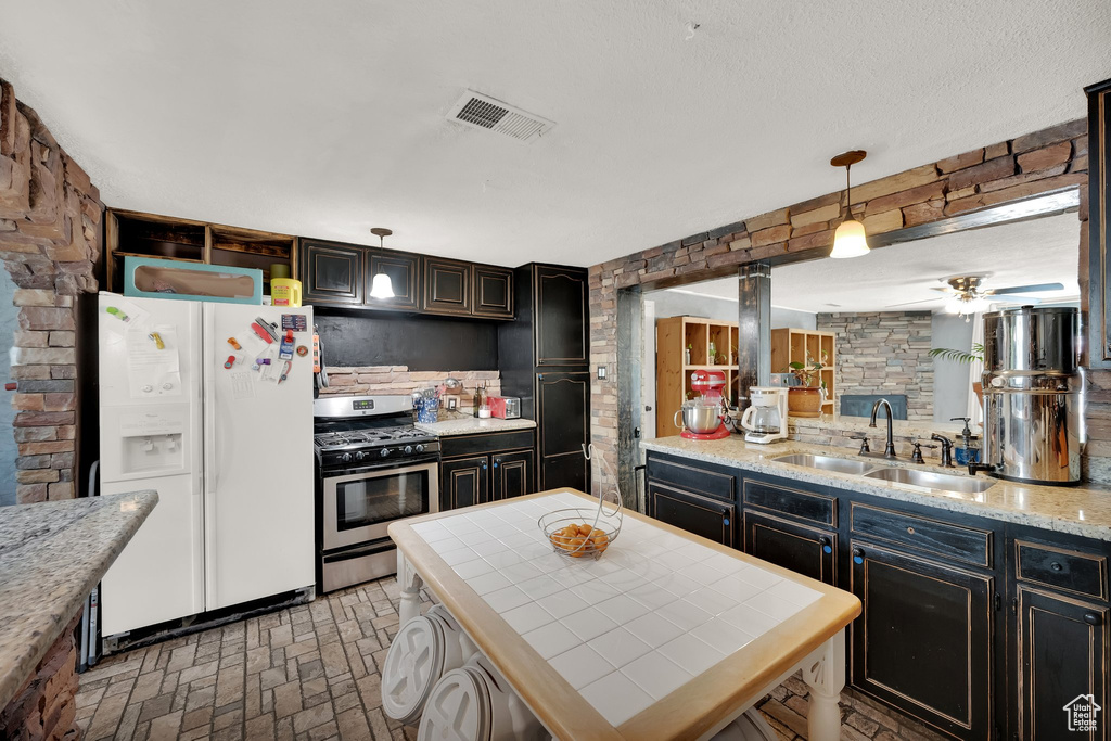 Kitchen featuring white fridge with ice dispenser, stainless steel range with gas cooktop, ceiling fan, brick patterned floors, and decorative light fixtures