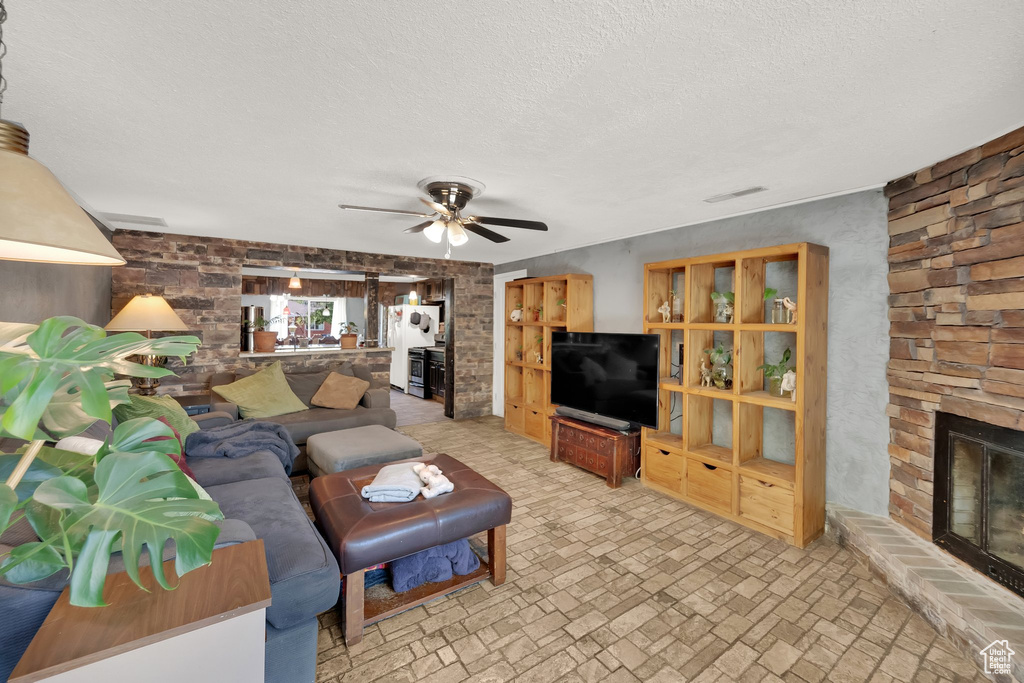 Living area with a stone fireplace, a ceiling fan, a textured ceiling, and brick patterned floors