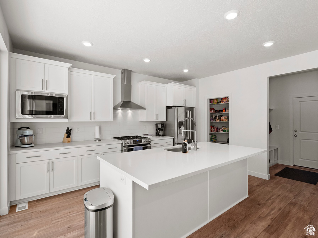 Kitchen featuring wall chimney exhaust hood, appliances with stainless steel finishes, a kitchen island with sink, decorative backsplash, and white cabinetry