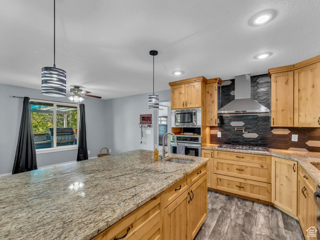 Kitchen featuring wall chimney range hood, appliances with stainless steel finishes, light brown cabinetry, a ceiling fan, and backsplash