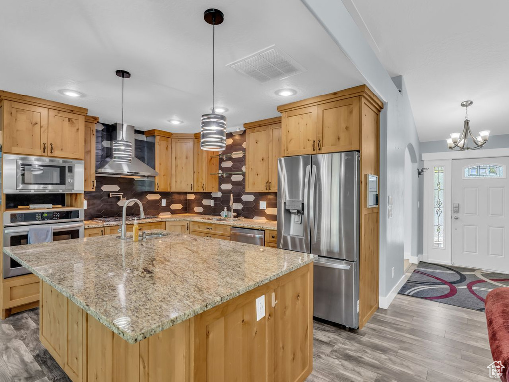 Kitchen with stainless steel appliances, wall chimney range hood, decorative backsplash, light wood-style flooring, and a chandelier