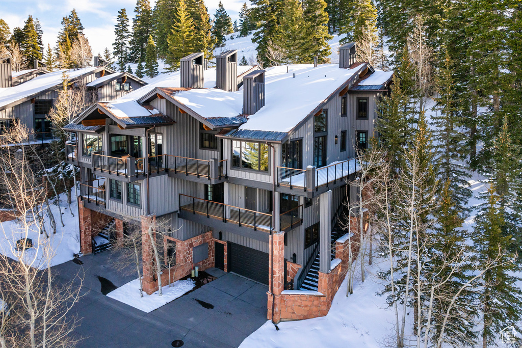 Snow covered back of property featuring a balcony, a garage, driveway, and a chimney