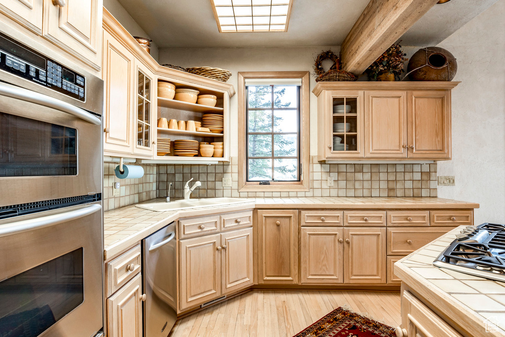 Kitchen with tile counters, appliances with stainless steel finishes, light wood finished floors, backsplash, and beam ceiling