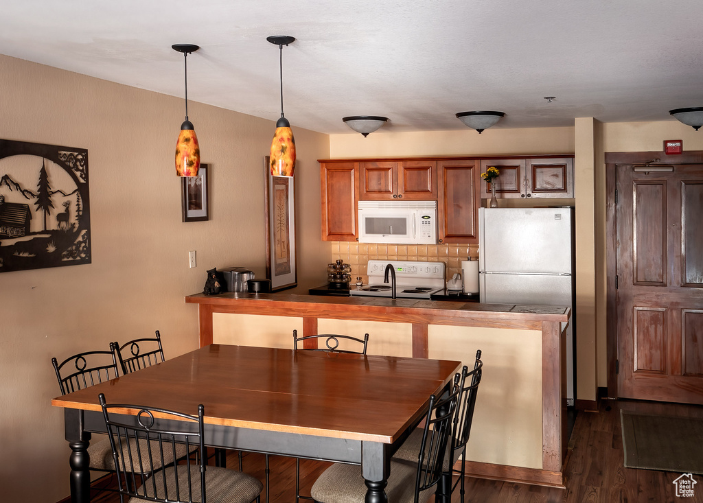 Kitchen featuring white appliances, dark countertops, decorative backsplash, dark wood-type flooring, and brown cabinetry
