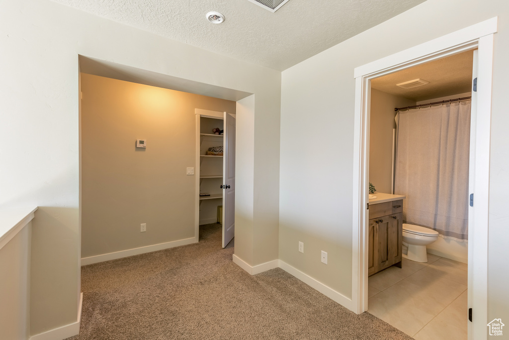 Unfurnished bedroom with light colored carpet, a walk in closet, ensuite bathroom, a textured ceiling, and light tile patterned flooring