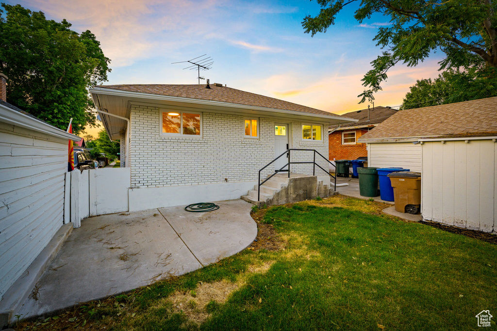 Back of property at dusk featuring brick siding, a yard, a patio area, an outdoor structure, and roof with shingles