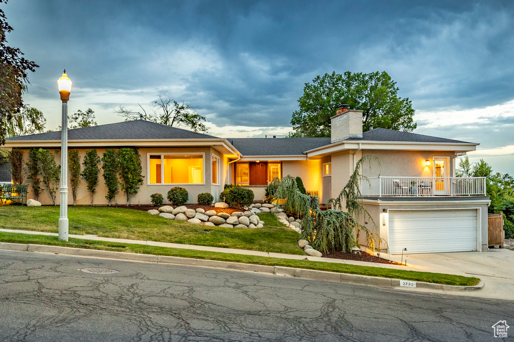 Ranch-style home featuring driveway, a chimney, a balcony, a garage, and a front yard