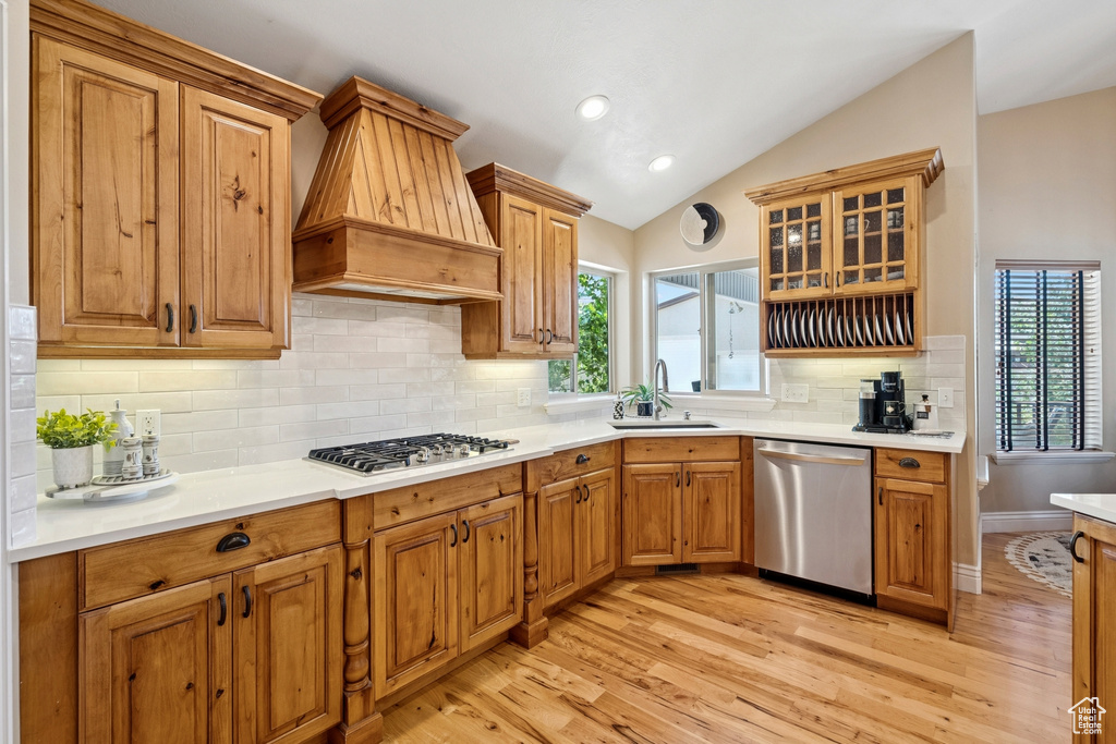 Kitchen with stainless steel appliances, custom exhaust hood, vaulted ceiling, light wood-style flooring, and plenty of natural light