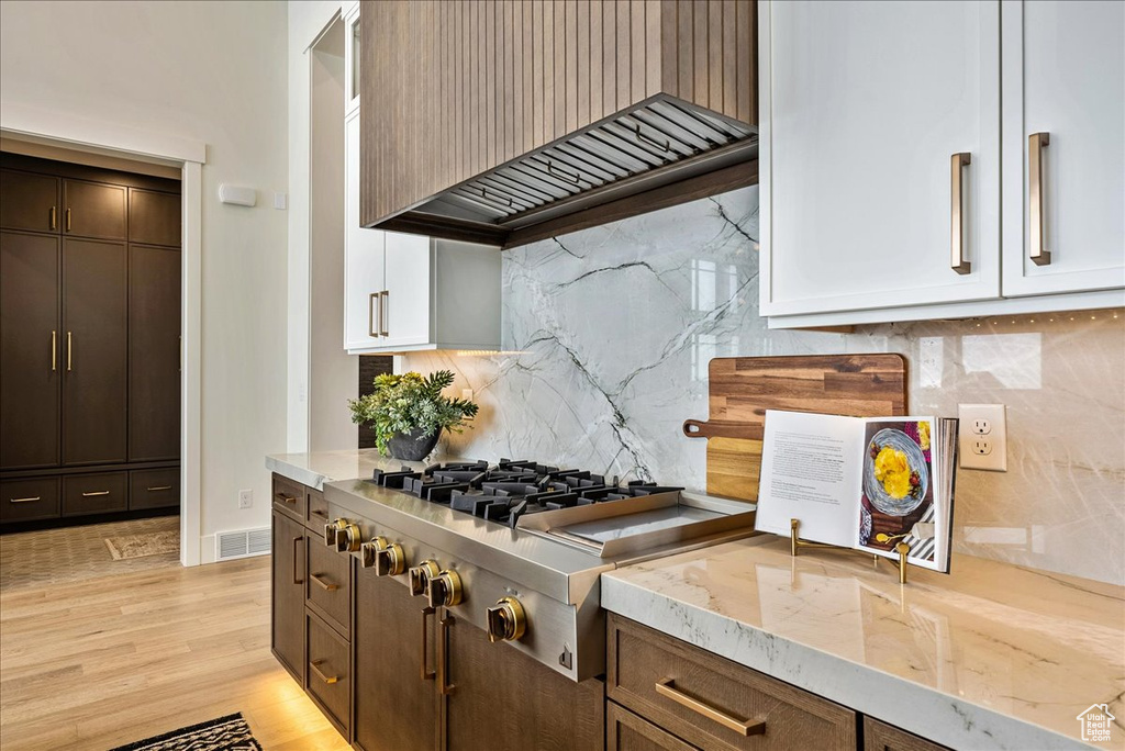 Kitchen with premium range hood, backsplash, light stone counters, light wood-style flooring, and stainless steel gas cooktop