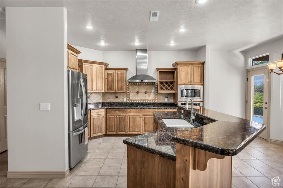 Kitchen with wall chimney range hood, stainless steel appliances, tasteful backsplash, open shelves, and an island with sink