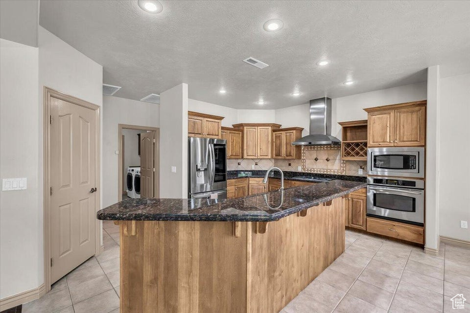 Kitchen with wall chimney range hood, stainless steel appliances, light tile patterned floors, a kitchen bar, and tasteful backsplash