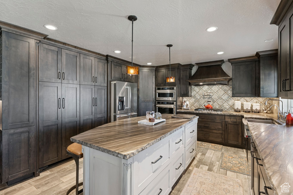 Kitchen featuring custom range hood, appliances with stainless steel finishes, a center island, light stone counters, and tasteful backsplash