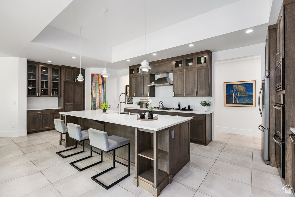 Kitchen featuring wall chimney exhaust hood, dark brown cabinetry, an island with sink, light tile patterned floors, and recessed lighting