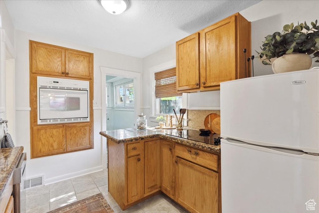 Kitchen featuring white appliances, dark stone countertops, a peninsula, a textured ceiling, and light tile patterned floors