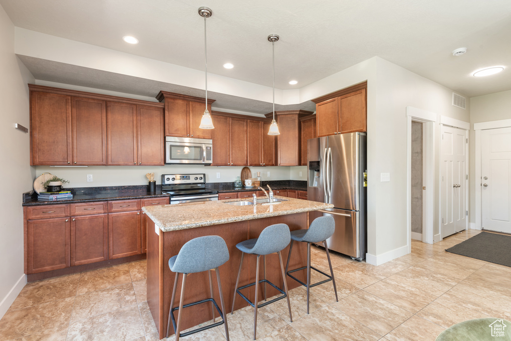 Kitchen with appliances with stainless steel finishes, brown cabinetry, dark stone countertops, a kitchen island with sink, and a kitchen breakfast bar