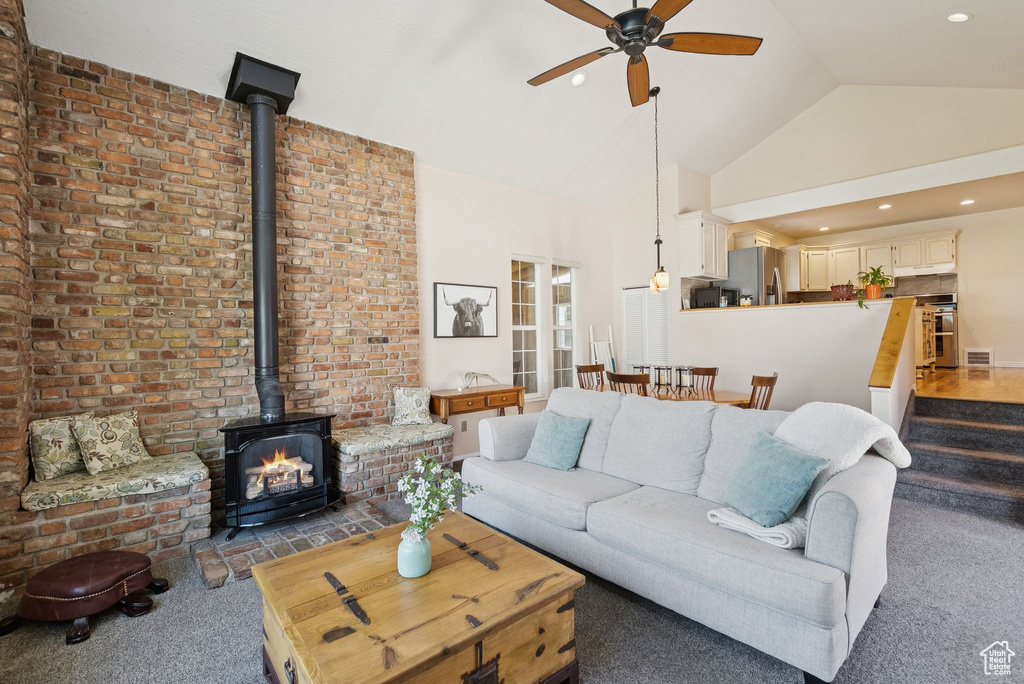 Living area with a wood stove, ceiling fan, carpet flooring, high vaulted ceiling, and brick wall